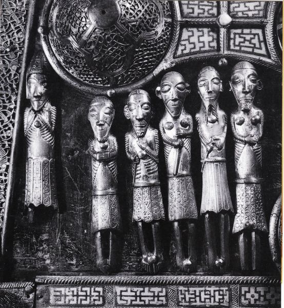 Detail van de beroemde reliekschrijn van Saint Manchán. Ze werd vervaardigd rond 1130 in klooster Clonmacnoise, bevat botresten, naar men aanneemt van Sint Manchán zelf. Ze bevindt zich tegenwoordig in de nabijgelegen parochiekerk van Boher. Er stonden ooit vijftig figuurtjes op. Daar zijn er nu nog elf van over op de onderste helft aan één van de zijkanten. Links in de hoek zien we een bisschop. Meer naar rechts, onder de knop en de arm van het kruis vijf mannen met baard, de ogen gesloten, de handen eerbiedig gevouwen. Twee van hen dragen een wapen. Nergens enige uitleg. Zou het gaan om Manchán? Zijn de figuurtjes monniken en soldaten? Omgekomen strijders van Diarmuid? Of zoeken we dan te ver?