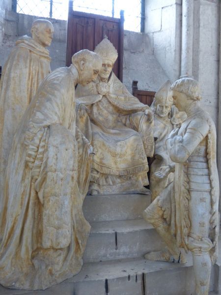 Gefingeerd proces van eerherstel voor Jeanne d'Arc Vanaf de 19e eeuw verandert de visie op Jeanne d'Arc. Enerzijds ziet men in haar het slachtoffer van kerkelijke heerszucht: zij is immers door kerkelijke hoogwaardigheidsbekleders ter dood veroordeeld. Het is de tijd dat het dogam van de pauselijke onfeilbaarheid wordt uitgeroepen! Na de door de Fransen verloren oorlog van 1870-1871 wordt zij symbool van vaderlandsliefde: zij staat voor de hoop van de vernederde Fransen en het verlangen naar eerherstel. Intussen vestigt de Kerk zelf de aandacht op haar heiligheid. Dat leidt ertoe dat zij in 1909 wordt zalig verklaard, en heilig in 1920. De afgebeelde personen verwijzen naar historische figuren, maar ze dragen de trekken van mensen uit de actualiteit van de kunstenaar. In Jeanne d'Arc zijn de trekken herkenbaar van zijn vrouw: Marie-Alice Laurent. De bisschop in het midden stelt Jean de Mailly voor (1425-1473), destijds bisschop van Noyon. Maar hij draagt de trekken van monseigneur Douais, bisschop van Beauvais ten tijde van de kunstenaar. De licht voorover gebogen man tegenover Jeanne verwijst naar de historische figuur van kanunnik Bouillé (1447-1476), deken van het kathedrale kapittel. Maar in hem herkent men Monseigneur Lagneaux (1888-1939), pastoor van de kathedrale kerk ten tijde van de kunstenaar. In de tijd tussen de beide Wereldoorlogen was het kunstwerk zo omstreden dat men het lange tijd niet in de kathedraal wilde hebben. [Gegevens ontleend aan een bijschrift in de kathedraal van Noyon]
