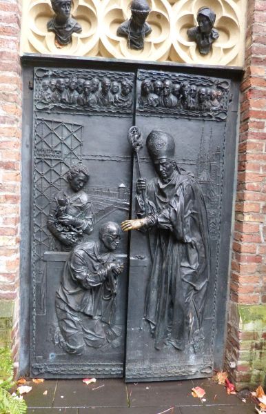 Priesterwijding Karl Leisner, 17 december 1944, door bisschop Piquet van Clermi nt-Ferrand. Links: Karl Leisner in werkkleding; achter hem houdt Zr Imma Mack alles gereed wat nodig is voor de wijding. Op het hek achter haar het - cynische - motto van de nazi's: 'Arbeit macht frei', Op de achtergrond: concentratiekamp Dachau. Rechts: strekt Bisschop Piquet zijn linkerhand uit, wit-goud glanzend door de vele aanrakingen, naar zijn wijdeling. Op de achtergrond de kathedraal van Clermont Ferrand. Boven hen de hoofden van 21 slachtoffers van de nazi's onder wie Anne Frank (1e van links op de rechter deur, en de jezuïetenpater Ruper Mayer (+ 1945; feest 3 november), helemaal rechts op de rechter deur. Boven hen de hoofden van links naar rechts: Stefanus, protomartelaar, Christus met gebroken boeien(!) en Lydia, eerste Europese persoon die het Christendom aanvaardde.
