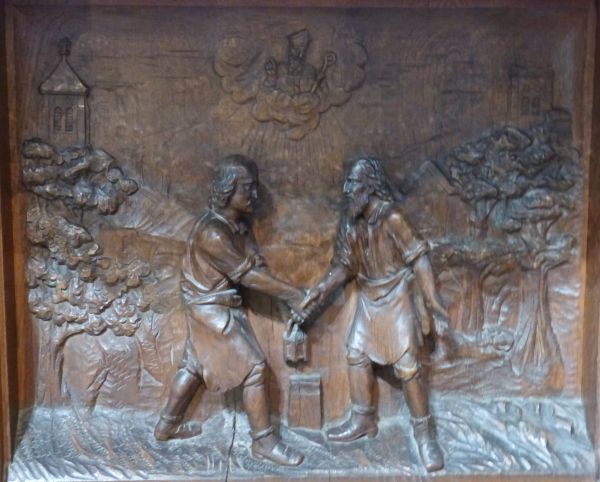 Sint Eligius (bisschop in de hemel!) verschijnt aan twee mannen met de opdracht een broederschap op te richten die zich zal bezighouden met het eerbiedig begraven van de doden. Zij bezegelen hun belofte met een onderlinge handdruk. ca 1920, houtreliëf.